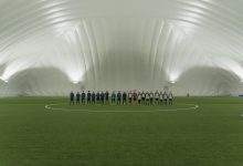 Duas equipes juvenis de futebol alinhadas em um campo de grama sintética sob a iluminação de um estádio coberto.