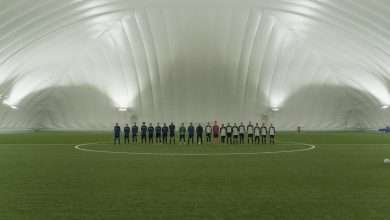 Duas equipes juvenis de futebol alinhadas em um campo de grama sintética sob a iluminação de um estádio coberto.