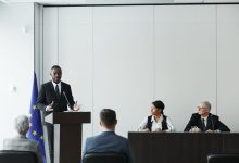 Profissionais participam de uma reunião institucional em ambiente interno, enquanto um palestrante fala ao microfone diante de um púlpito.