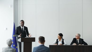 Profissionais participam de uma reunião institucional em ambiente interno, enquanto um palestrante fala ao microfone diante de um púlpito.