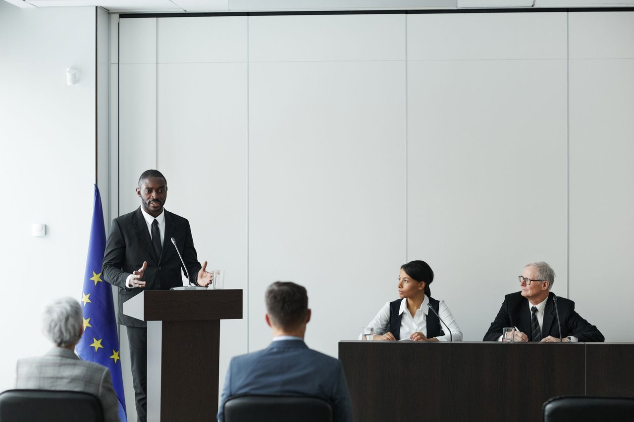 Profissionais participam de uma reunião institucional em ambiente interno, enquanto um palestrante fala ao microfone diante de um púlpito.